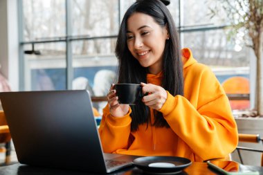 Kafede kahve içerken dizüstü bilgisayarla çalışan gülümseyen Asyalı kadın.