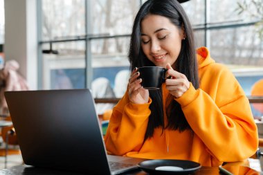 Kafede kahve içerken dizüstü bilgisayarla çalışan gülümseyen Asyalı kadın.