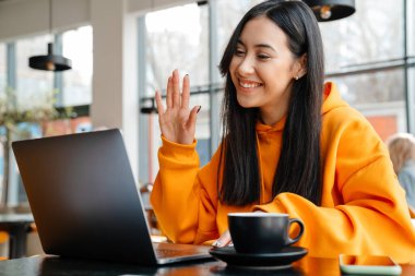 Gülümseyen Asyalı kadın kafede otururken diz üstü bilgisayar kullanıyor.