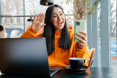 Kafede dizüstü bilgisayarla çalışırken cep telefonu kullanan gülümseyen Asyalı kadın.