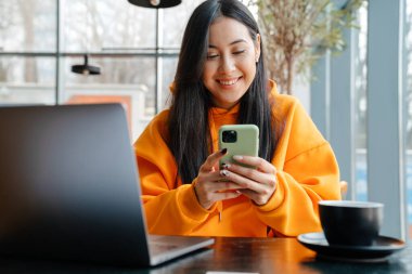 Kafede dizüstü bilgisayarla çalışırken cep telefonu kullanan gülümseyen Asyalı kadın.