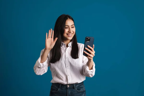 Happy beautiful asian girl waving hand and using smartphone isolated over blue background