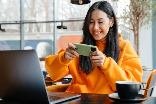 Uśmiechnięta Azjatka Gra Online Telefon Komórkowy Podczas Pracy Laptopem Kawiarni — Zdjęcie stockowe