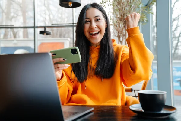 Heyecanlı Asyalı kadın internette cep telefonuyla oyun oynuyor ve kafede birinci sınıf jest yapıyor.