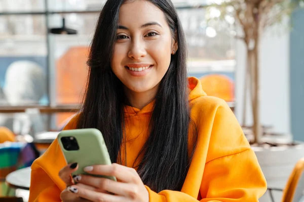 Uśmiechnięta Azjatka Korzystająca Telefonu Komórkowego Podczas Siedzenia Kawiarni — Zdjęcie stockowe
