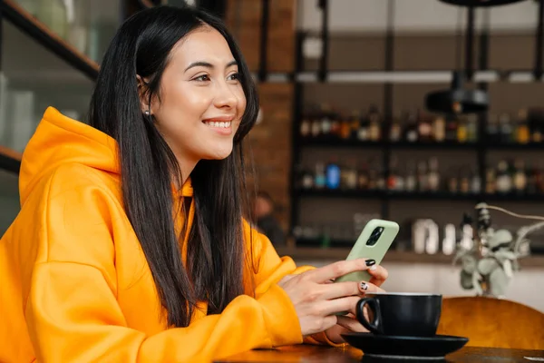 Uśmiechnięta Azjatka Korzystająca Telefonu Komórkowego Podczas Siedzenia Kawiarni — Zdjęcie stockowe