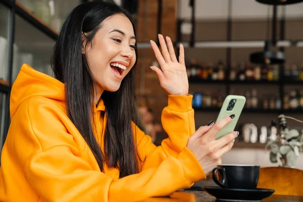 Uśmiechnięta Azjatka Gestykulująca Korzystająca Telefonu Komórkowego Siedząc Kawiarni — Zdjęcie stockowe