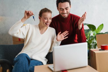 Yeni evde dizüstü bilgisayarı olan gülümseyen bir çift, anahtarları gösteriyor.