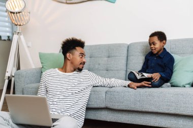 Odaklanmış Afro-Amerikan baba-oğul kitap okuyor ve evde otururken dizüstü bilgisayar kullanıyor.