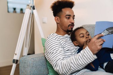 Odaklanmış Afro-Amerikan baba ve oğlu evdeki kanepede dinlenirken kitap okuyorlar.