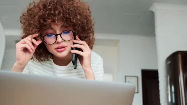 家のノートパソコンの前に座っていると 電話で話している眼鏡の中の笑顔の巻き毛の女の子のクローズアップ — ストック動画