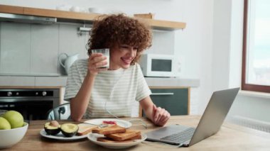Kablolu kulaklıklı olumlu kıvırcık saçlı bir kız bilgisayardaki video görüşmesinde konuşuyor ve mutfakta reçelli tost yiyor.