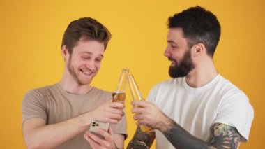 İki memnun adam, ellerinde akıllı telefonlarıyla stüdyodaki sarı duvarın üzerinde duran bira şişelerini tutuyorlar.
