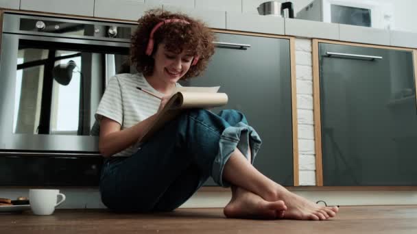 床に座っている間に何かを描くヘッドフォンの笑いの巻き毛の女の子 — ストック動画