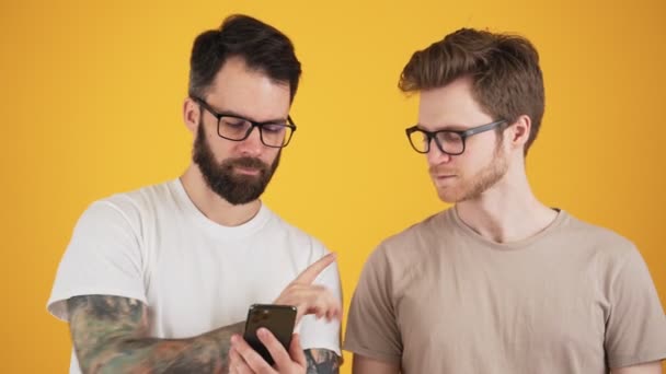 Два Симпатичных Парня Очках Обсуждают Держа Смартфон Изоляции Над Жёлтой — стоковое видео