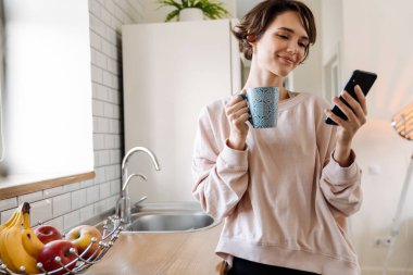 Ev mutfağında kahve içerken cep telefonu kullanan neşeli, güzel bir kız.