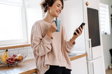 Ev mutfağında kahve içerken cep telefonu kullanan neşeli, güzel bir kız.