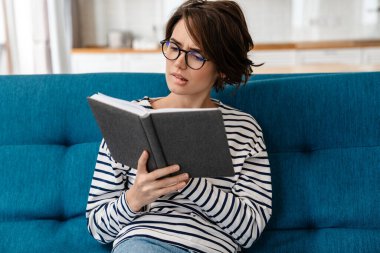 Gözlüklü düşünceli güzel kadın evde kanepede otururken kitap okuyor.
