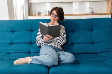 Gözlüklü düşünceli güzel kadın evde kanepede otururken kitap okuyor.