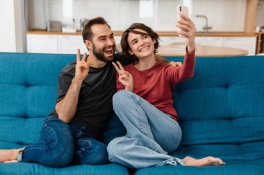Mutlu genç çift evde cep telefonuyla selfie çekerken barış işareti gösteriyor.