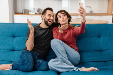 Mutlu genç çift evde cep telefonuyla selfie çekerken havada öpüşüyor.