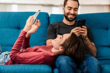 Mutlu genç çift evde kanepede dinlenirken gülümsüyor ve cep telefonu kullanıyor.