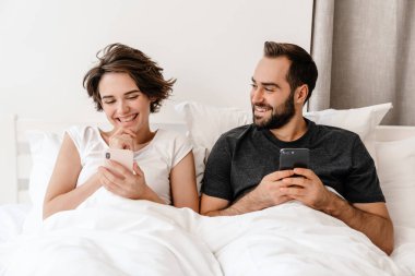 Mutlu genç çift evde yatarken gülümsüyor ve cep telefonu kullanıyor.
