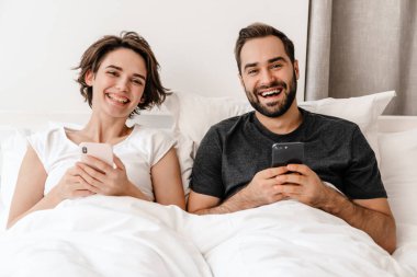 Mutlu genç çift evde yatarken gülüyor ve cep telefonu kullanıyor.
