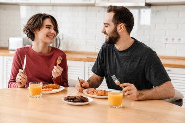 Mutlu genç çift evde kahvaltı ederken gülümsüyor ve konuşuyor.