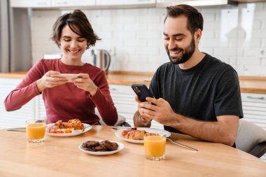 Evde rahat bir mutfakta kahvaltı ederken cep telefonuyla fotoğraf çeken beyaz bir çiftin portresi.