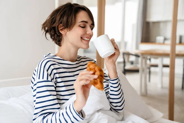快乐美丽的女人坐在沙发上 手里拿着咖啡和羊角面包 — 图库照片