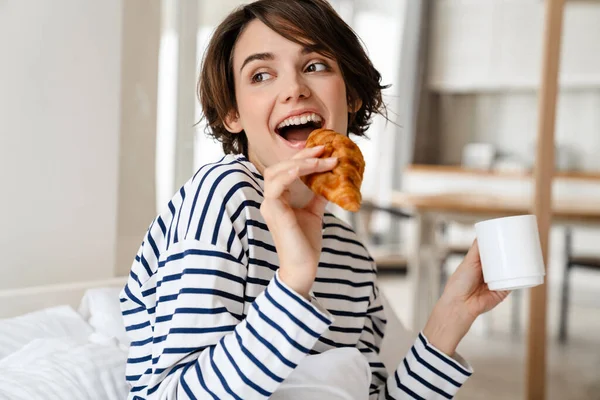 Mulher Bonita Feliz Segurando Xícara Café Croissant Enquanto Sentado Sofá — Fotografia de Stock