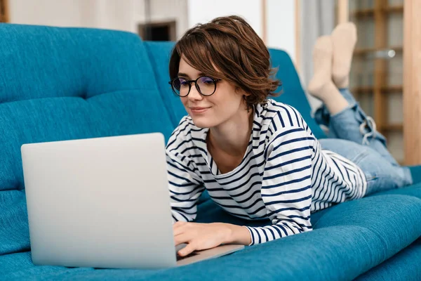 Mulher Bonita Feliz Óculos Usando Laptop Enquanto Deitado Sofá Casa — Fotografia de Stock