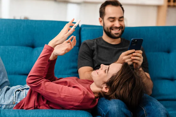Feliz Joven Pareja Sonriendo Usando Teléfonos Móviles Mientras Descansa Sofá —  Fotos de Stock