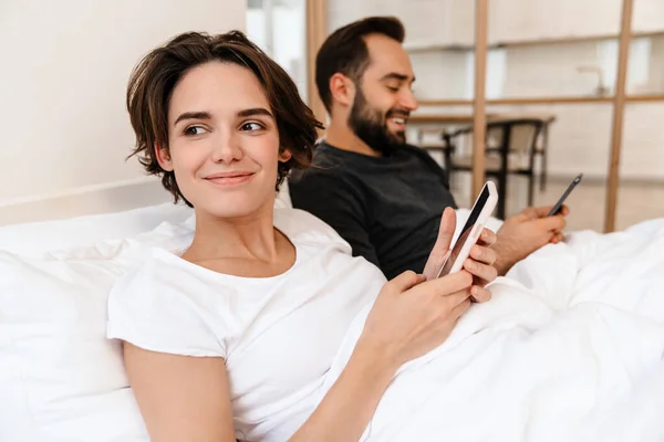 Glückliches Junges Paar Lächelt Und Benutzt Mobiltelefone Während Hause Bett — Stockfoto