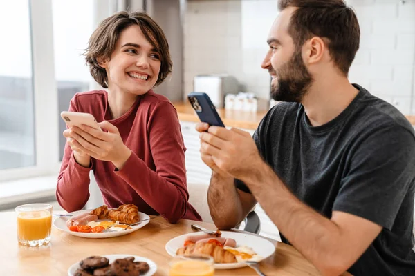 Feliz Hermosa Pareja Usando Teléfonos Celulares Mientras Desayuna Casa — Foto de Stock