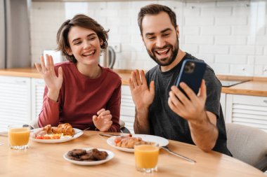 Neşeli çift evde kahvaltı ederken el sallıyor ve cep telefonu kullanıyor.
