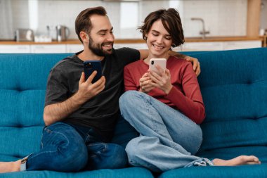 Mutlu genç çift evde koltukta otururken gülümsüyor ve cep telefonu kullanıyor.