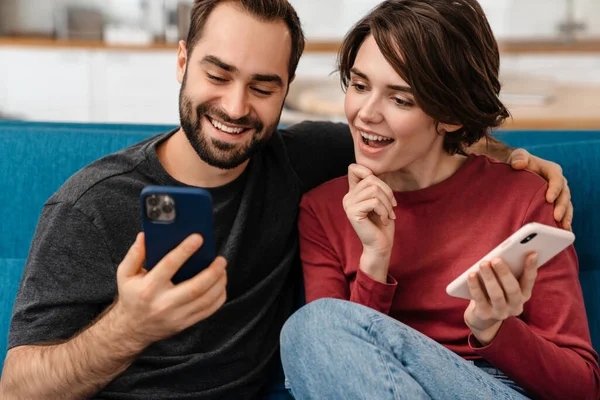 Podekscytowana Młoda Para Uśmiechnięta Korzystająca Telefonów Komórkowych Siedząc Kanapie Domu — Zdjęcie stockowe