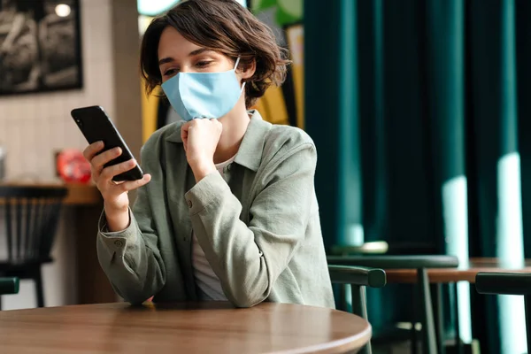 Menina Feliz Máscara Protetora Usando Celular Enquanto Sentado Café Dentro — Fotografia de Stock