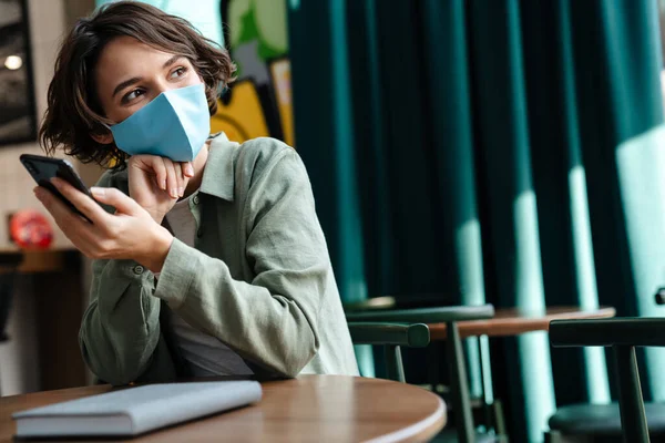Menina Feliz Máscara Protetora Usando Celular Enquanto Sentado Café Dentro — Fotografia de Stock