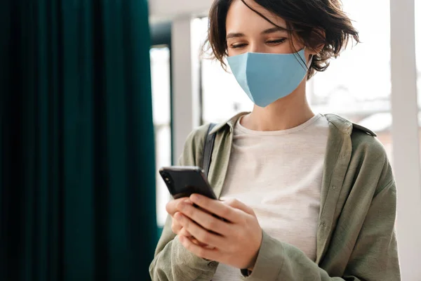 Happy Girl Protective Mask Using Cellphone While Standing Indoors — Stock Photo, Image