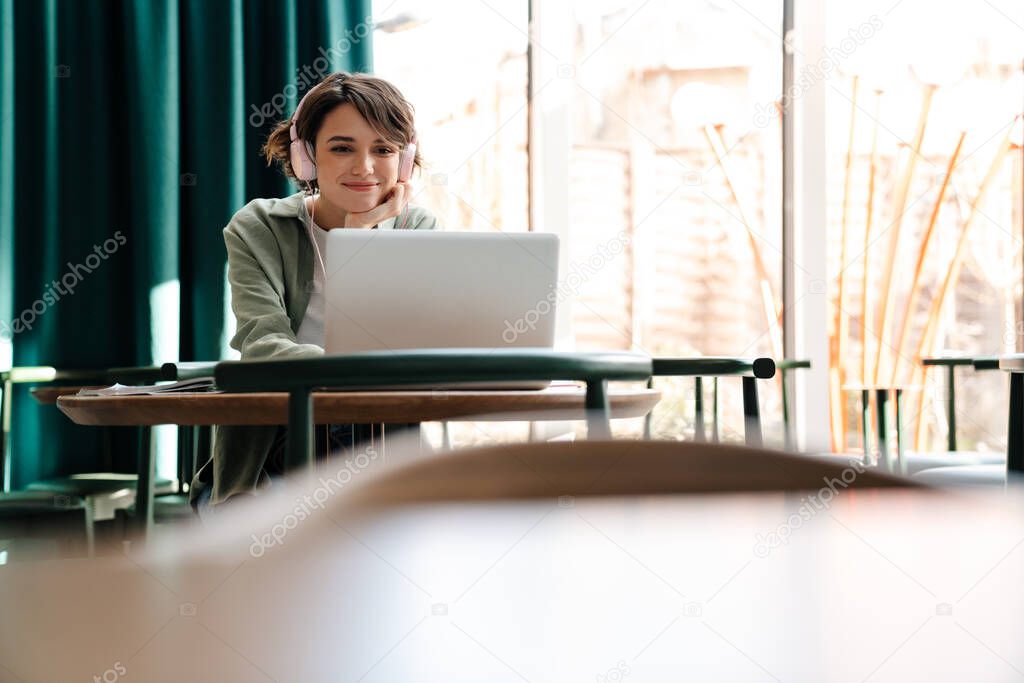 Smiling caucasian girl using headphones and laptop while sitting in cafe indoors
