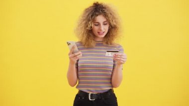 Sarışın güzel bir kadın, elinde sarı duvarın üzerinde duran bir kredi kartı tutarken akıllı telefonunu kullanıyor.