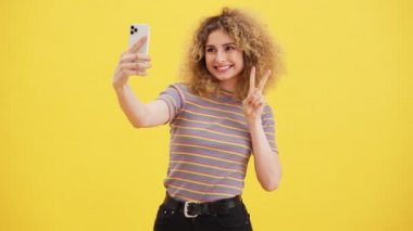 Pozitif sarışın bir kadın, stüdyodaki sarı duvarın üzerinde tek başına dururken akıllı telefonunu kullanarak selfie çekiyor.