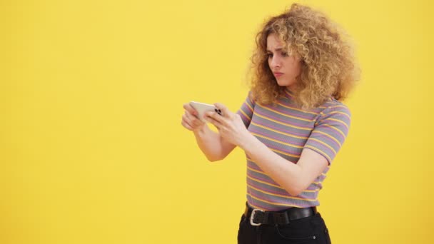 一位专心致志的金发女人正在用她的智能手机玩游戏 但以失败告终 站在工作室的黄墙上 — 图库视频影像