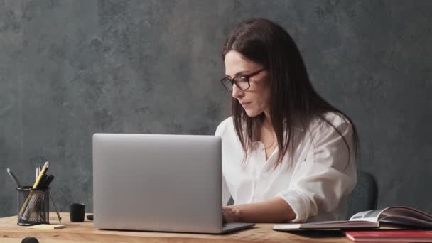 眼鏡をかけたビジネスウーマンがオフィスのテーブルに座りながらノートパソコンや書類を操作している — ストック動画