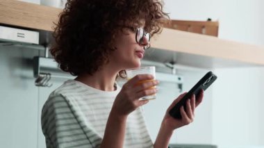 Gözlüklü, kendine güvenen, kıvırcık saçlı bir kızın telefondaki bandı yapıştırması ve mutfakta bir bardak tutması.