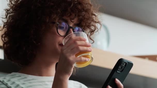 携帯電話を見て キッチンでジュースを飲む眼鏡で肯定的な巻き毛の女の子のクローズアップビュー — ストック動画