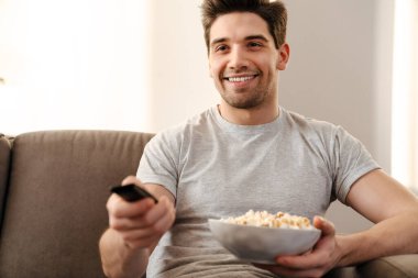 Mutlu adam televizyon seyrediyor ve evde kanepede patlamış mısır yiyor.